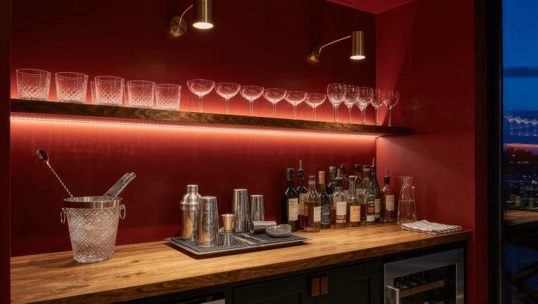 A compact home bar with distinct stations for glassware, ice, tools, and bottles under LED shelf strips and brass pendants at dusk.