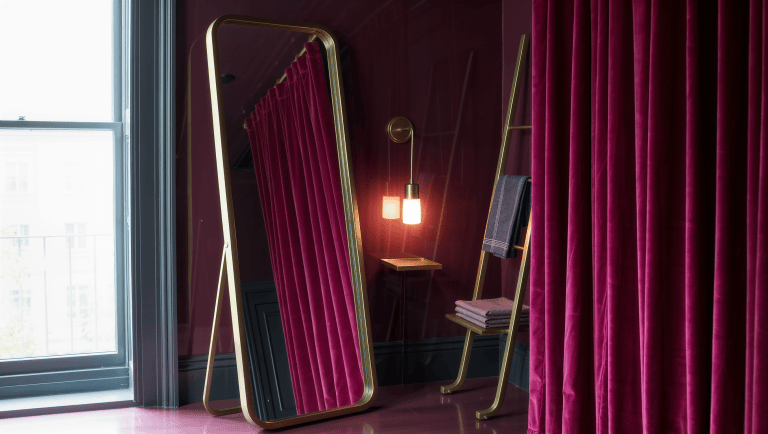 A freestanding brass-framed leaning mirror propped against a lacquered plum wall beside a tension-rod velvet curtain, no wall attachments visible.