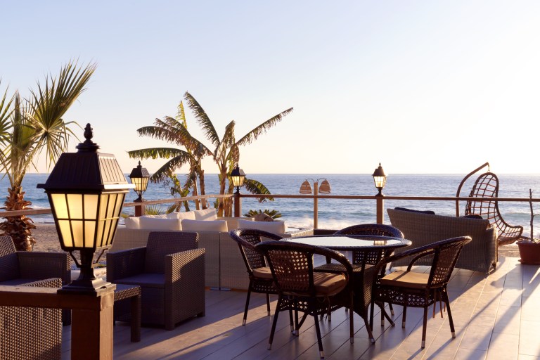 A rooftop terrace glows at dusk, its wicker table and chairs lit by an iron lantern beside silhouetted palms and a darkening sea horizon.