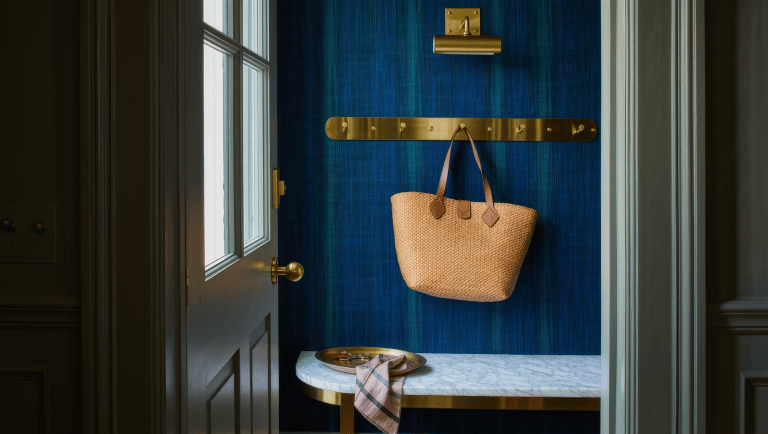 A marble-topped entry bench beneath brass wall hooks holds a woven tote and a tray for keys beside an open door.