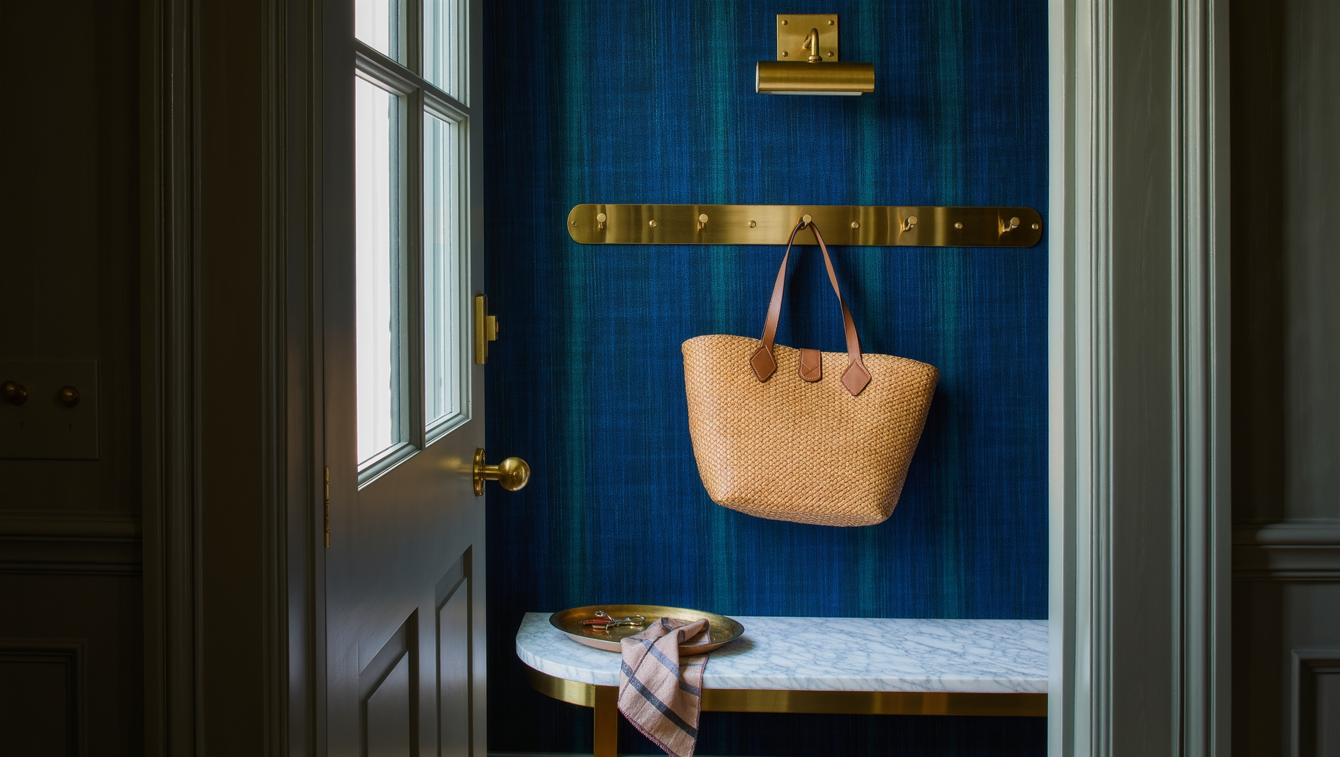 A marble-topped entry bench beneath brass wall hooks holds a woven tote and a tray for keys beside an open door.