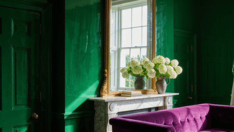 Gilt mirror above a marble mantel reflecting a bright window and doubling the daylight across a plum sofa and deep green wall.