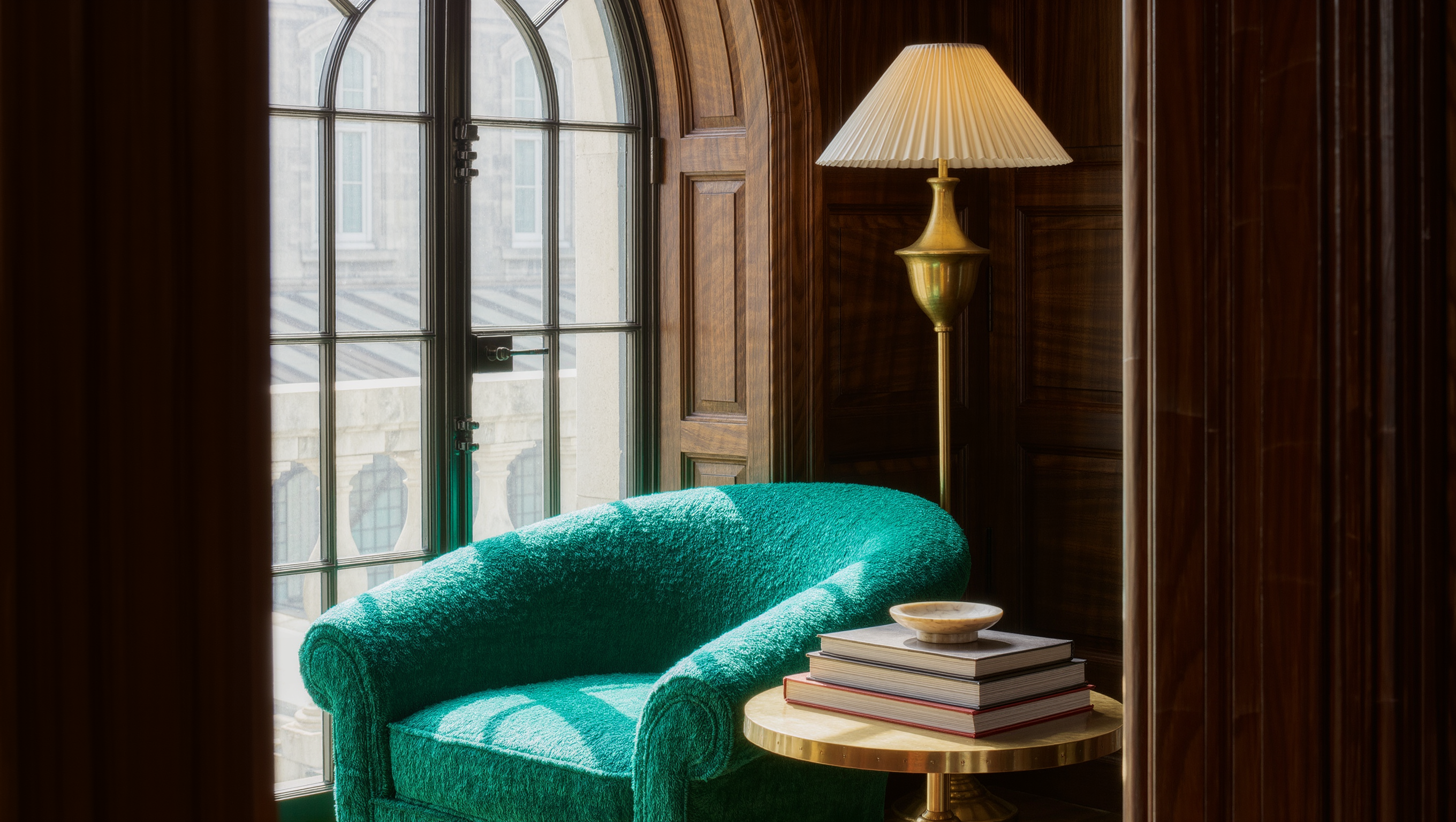 A curved emerald armchair and brass floor lamp form a reading corner beside a tall window.