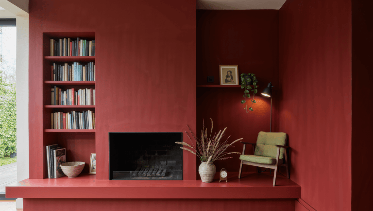 An oxblood-painted chimney breast flanked by two alcoves, one fitted with a bookcase and the other holding a small reading chair.