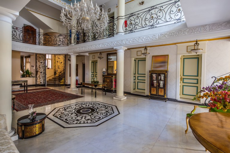 A grand marble-floored hall with freestanding stone columns rising to an ornate gilded ceiling, a wrought-iron balustrade above, and no furniture placed against them.