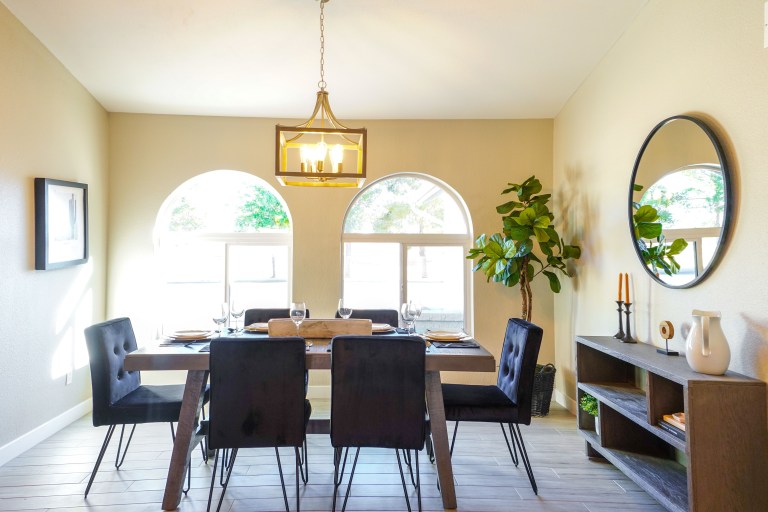 A dark wood dining table with black upholstered chairs sits beneath a brass chandelier between arched windows.