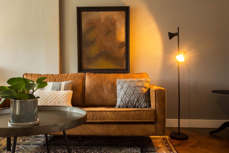 A cognac leather sofa layered with mixed-pattern pillows sits beside a small round accent table, framed art, and a brass floor lamp on a patterned rug.