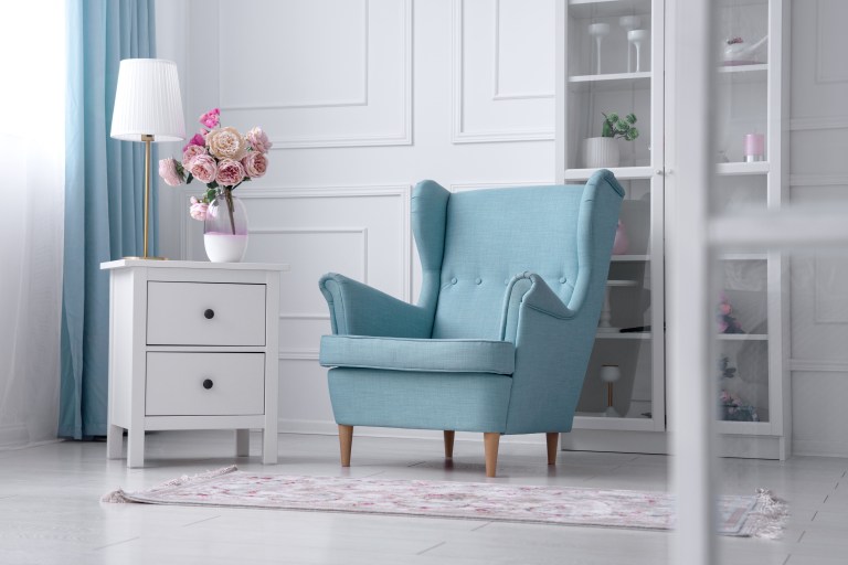 A sapphire-blue wingback armchair angled beside a mirrored dresser with fresh flowers, in a bright, classically trimmed room.