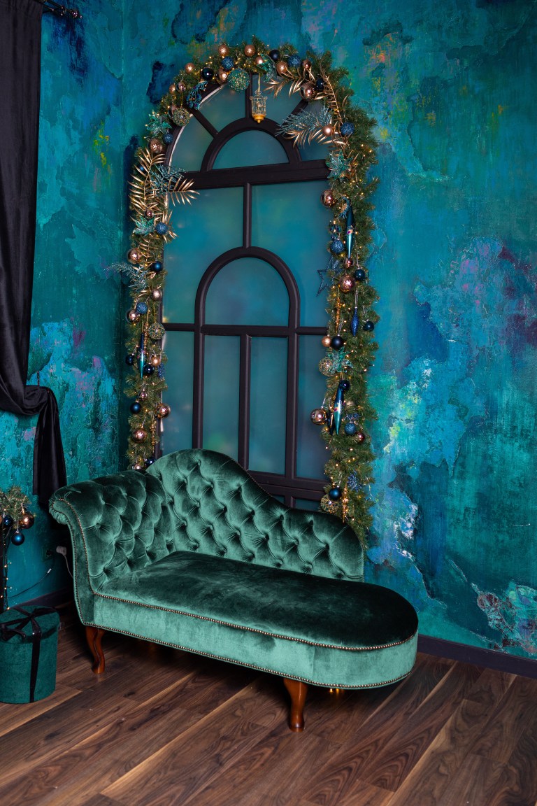 A tufted emerald velvet chaise longue set against a richly textured jewel-toned wall, beneath an ornate arched mirror framed in gilded foliage.