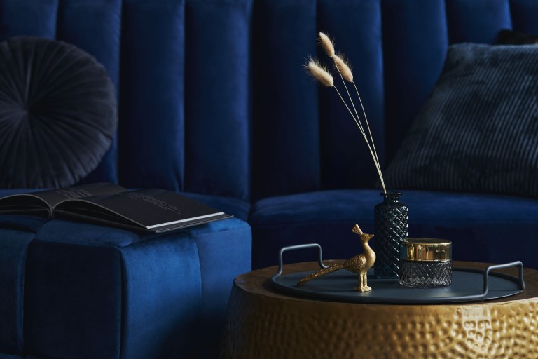 A navy channel-tufted velvet sofa corner with an open book, beside a round hammered brass side table holding a dark ceramic vase of dried branches.
