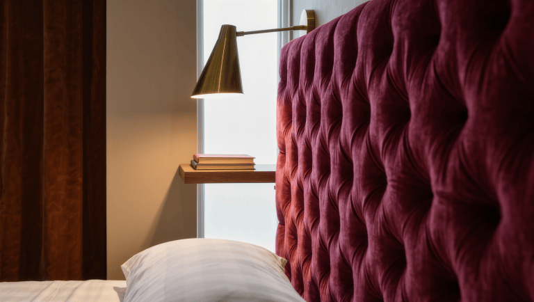 A tufted burgundy velvet bed corner with a floating brass reading light along a narrow wall