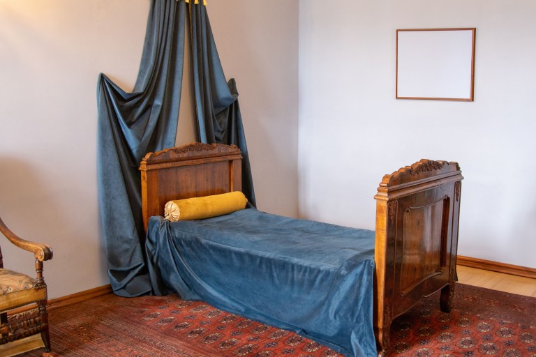 A carved wood bed dressed in sapphire velvet with a matching teal canopy drape, set along a narrow wall over a richly patterned rug.