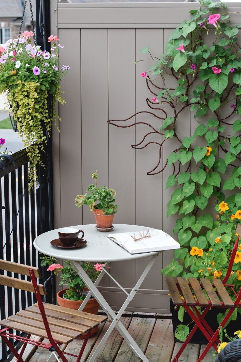 A small apartment balcony with a white bistro table and folding chairs surrounded by climbing vines and blooming potted flowers, a coffee cup and open book on the table.