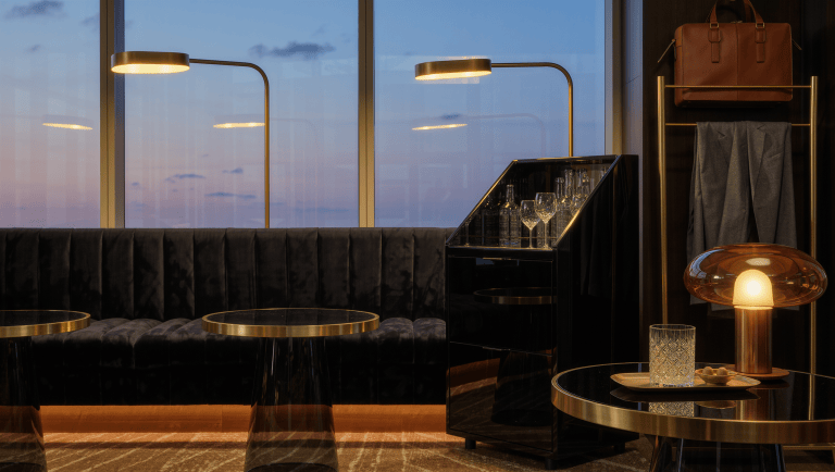 A hospitality-style lounge with a charcoal velvet banquette, black-lacquer drinks cabinet, brass rail with a leather bag, and warm lamps at dusk.