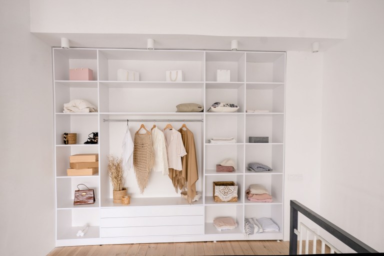 A floor-to-ceiling white built-in wardrobe with integrated lighting, its open shelves and cubbies holding neatly folded linens, garments hung on a center rod, and woven storage baskets.