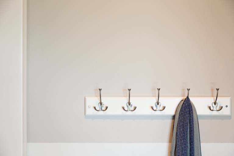 A row of polished wall hooks mounted on a plain painted wall, with a single dark wool coat hanging from one hook.