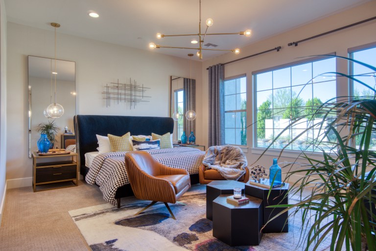 A polished bedroom vignette with a navy velvet headboard, warm bedside lighting, a leather accent chair, and brass-framed decor reflected in a floor mirror.