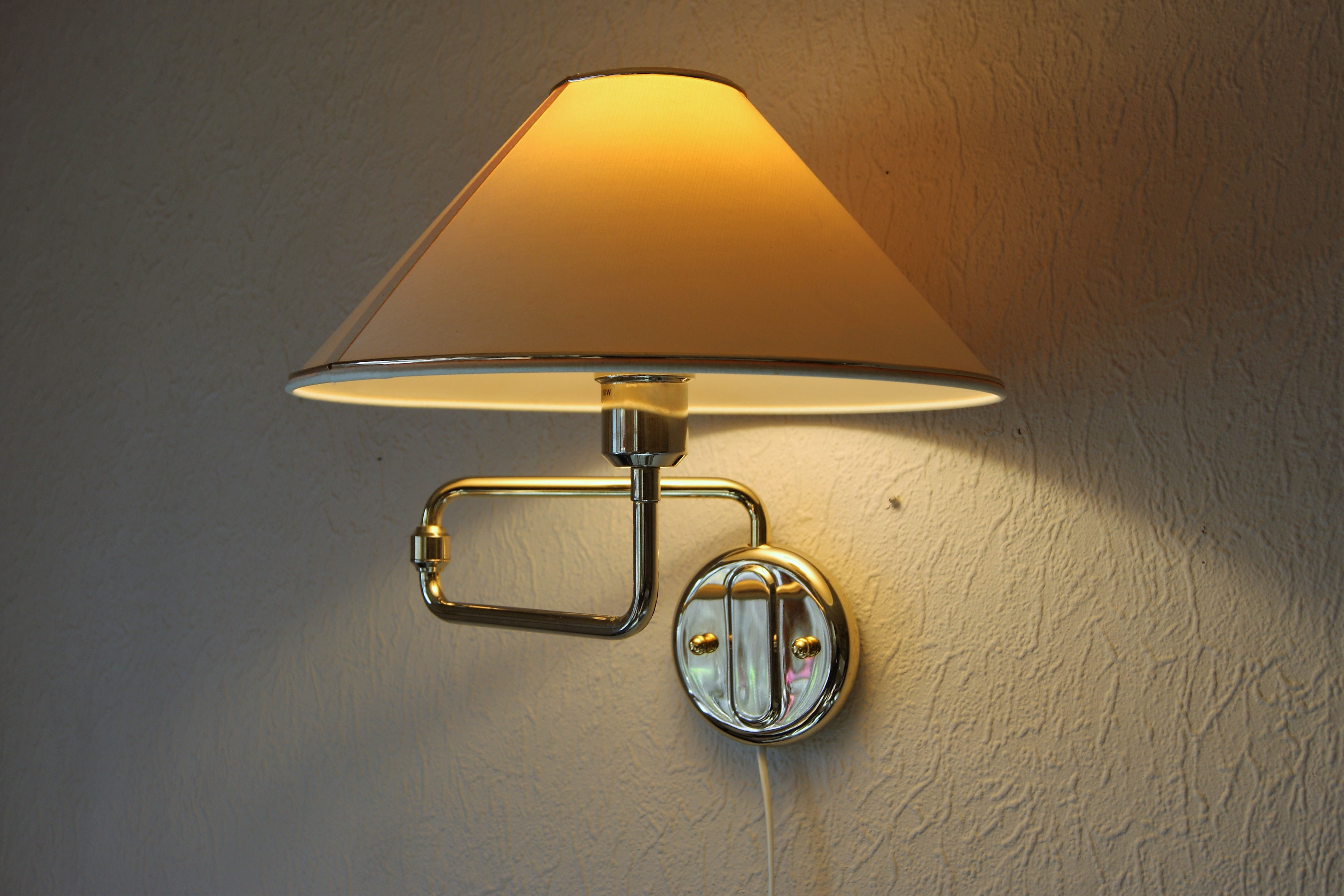 A polished brass swing-arm wall sconce with a cream fabric shade, mounted directly on the wall with its arm extended out to cast warm light over the surface below.