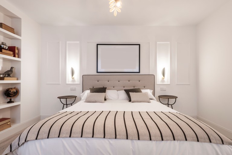 A built-in bed alcove flanked by open bookshelves and recessed wall sconces, with a tufted taupe headboard and striped linens.