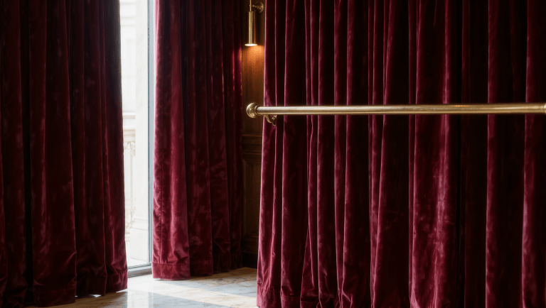 A heavy jewel-toned burgundy velvet curtain hanging fully closed against a tall window, its lined fabric pooling slightly on marble to block drafts.