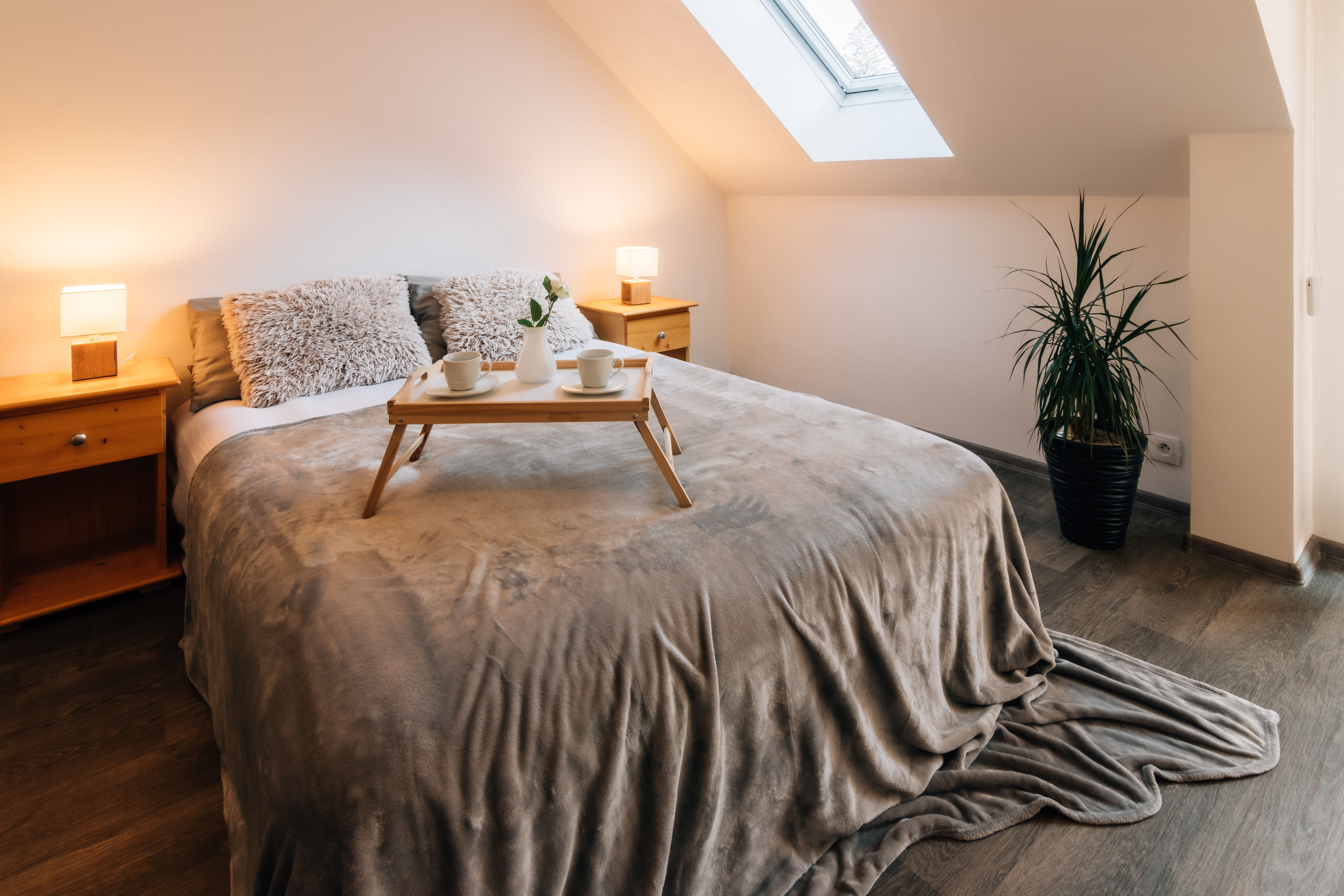 An attic bedroom tucked beneath a sloped ceiling with a skylight, dressed in warm gray linens with a wood breakfast tray and a leafy plant in the corner.
