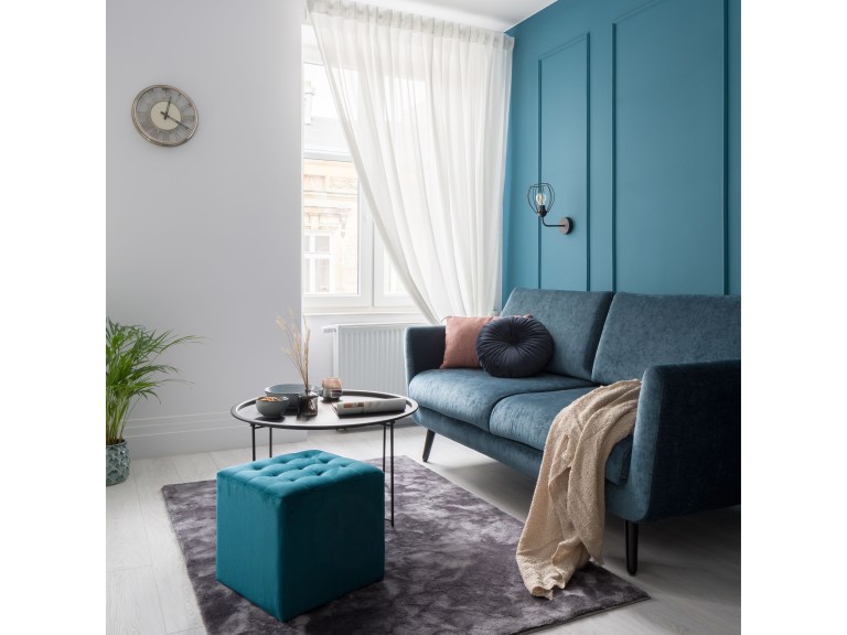 A jewel-toned teal living room with deep blue paneled wall, a velvet sofa, and a matching velvet ottoman styled with brass sconces and layered textiles.