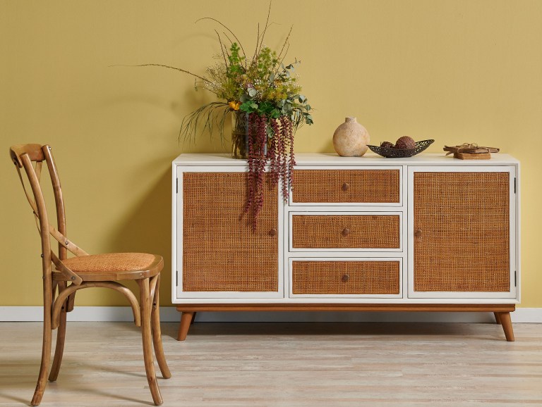 A white and rattan sideboard stands against a rich mustard-yellow wall, styled with a trailing plant and ceramic accessories, with a woven-seat chair beside it.