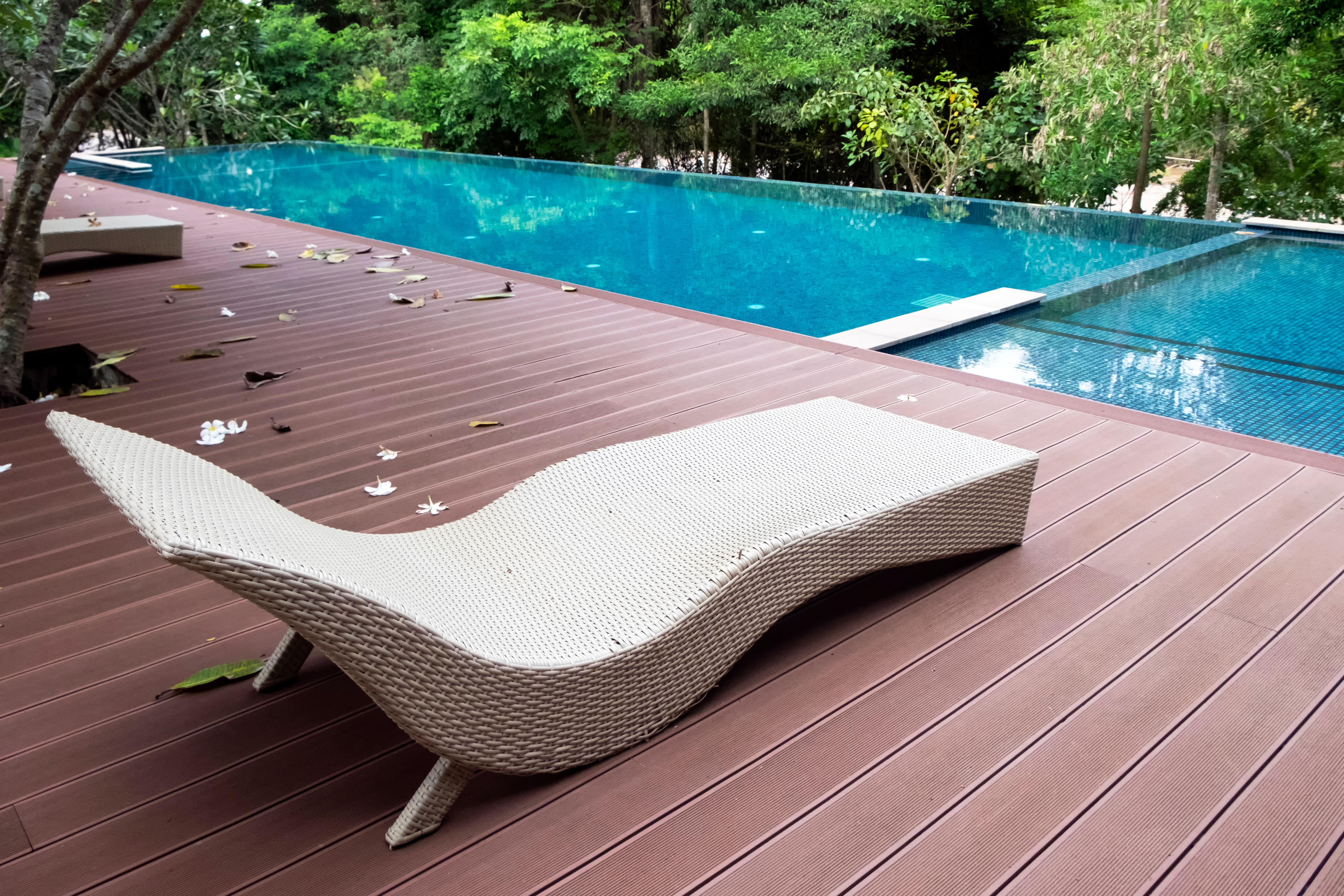 A cream wicker chaise lounge sits on a private wood deck beside a pool, screened by dense trees.