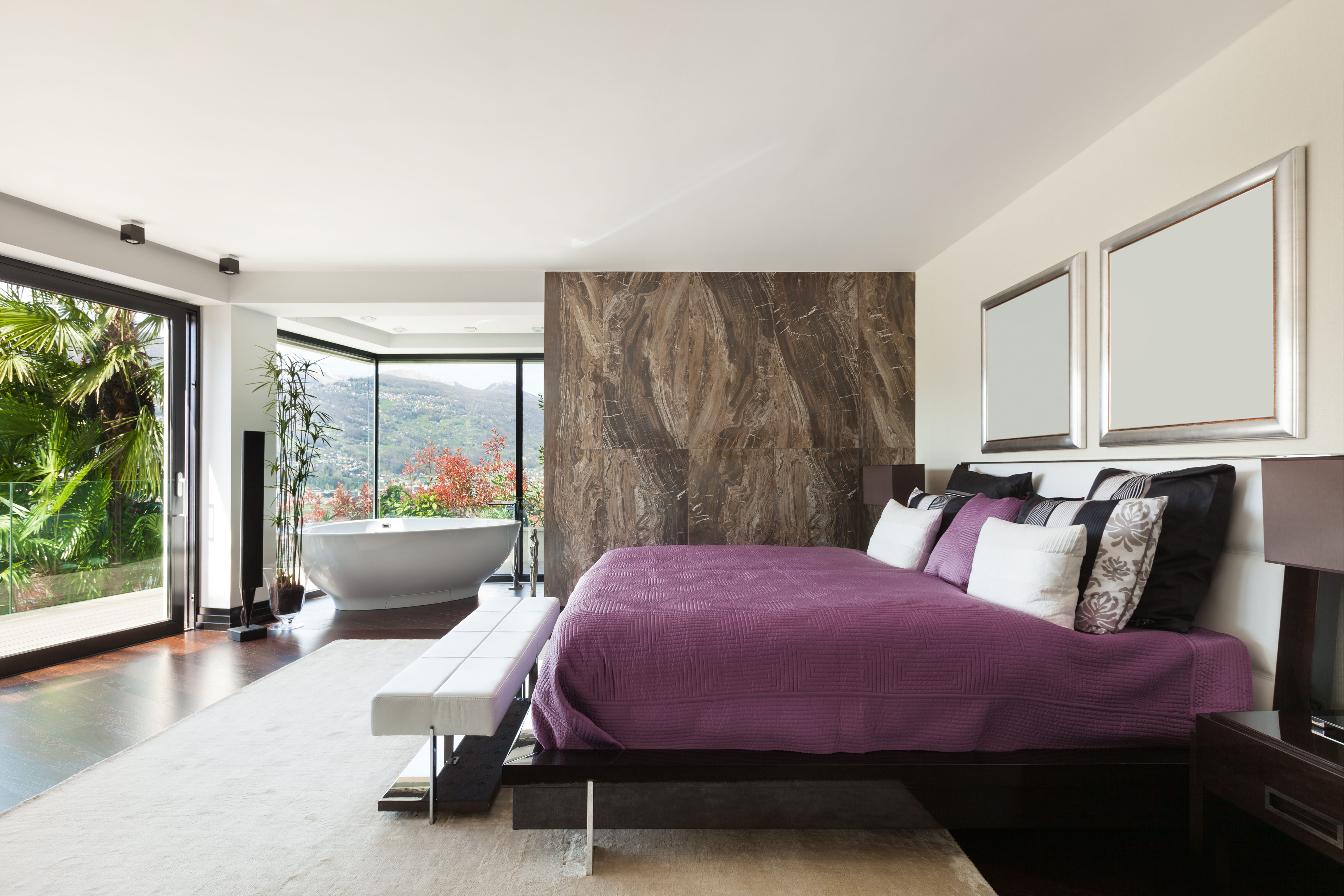 A plum-draped bed sits before a dramatic marble accent wall, with a low white bench positioned at its foot and a freestanding tub visible beyond glass doors.