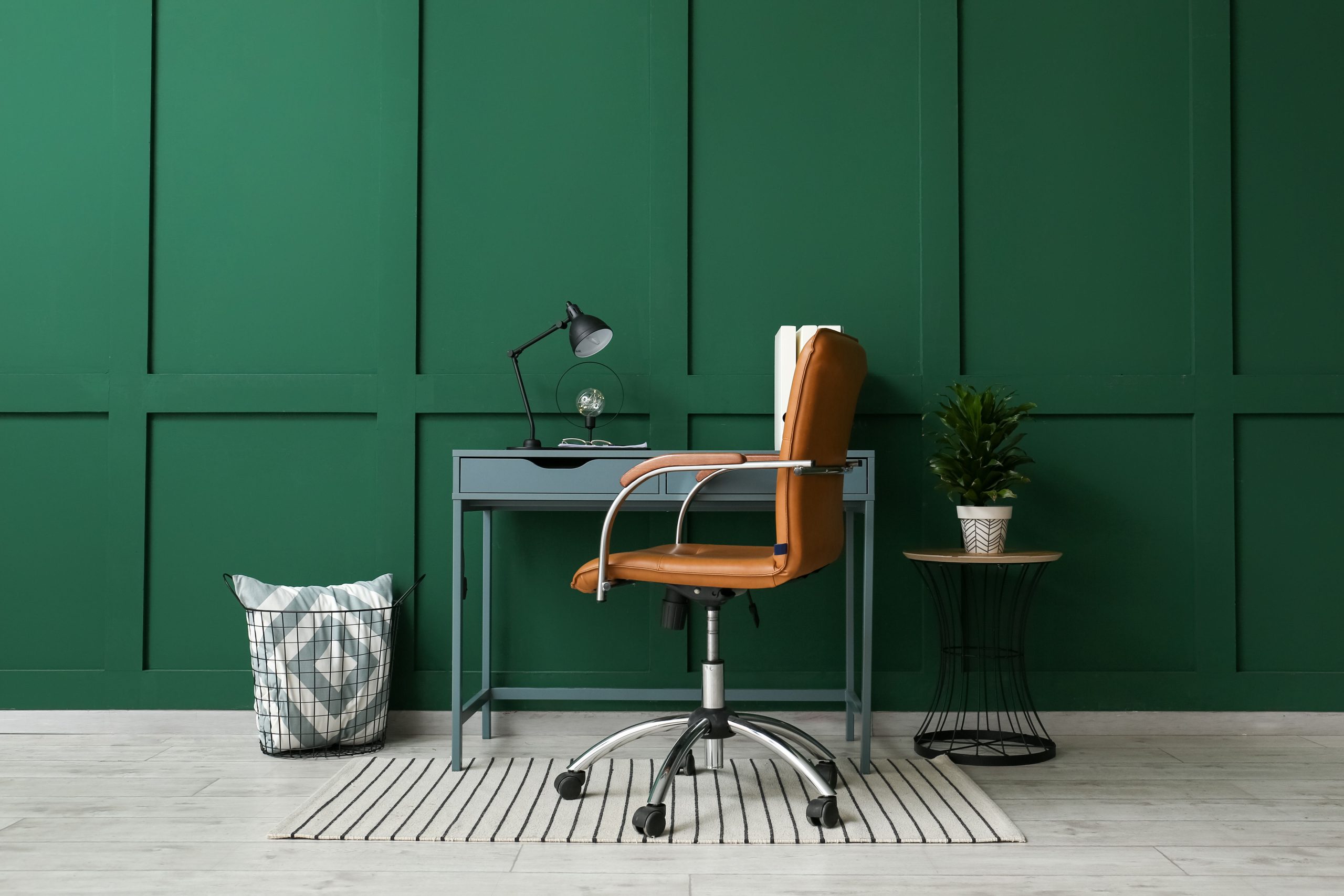 A slim desk with a brass lamp and cognac leather chair sits against an emerald green paneled wall.