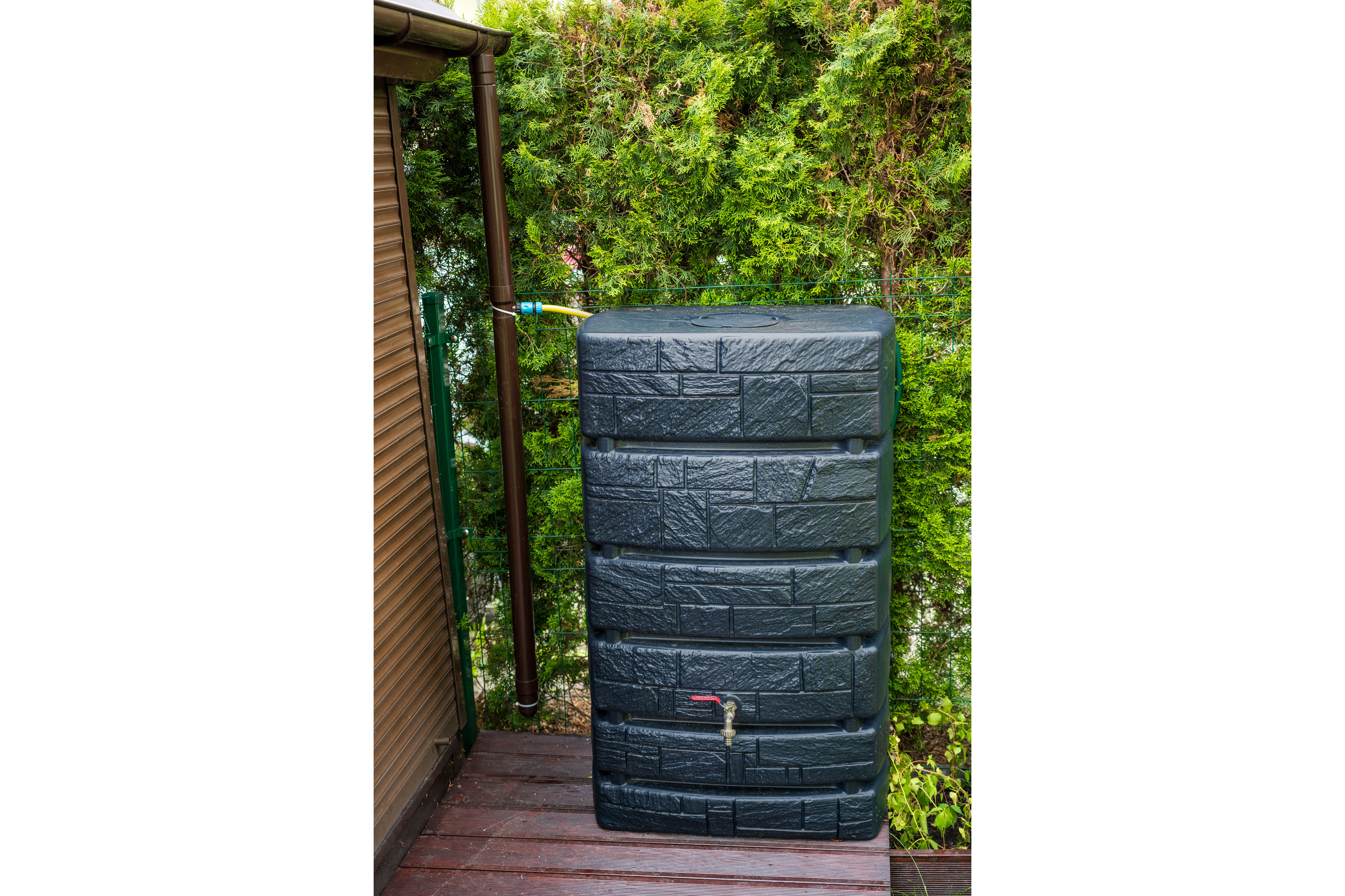 A tall, modern stone-textured rainwater tank fitted with a spigot, positioned neatly on a wood deck beside a dense green hedge.