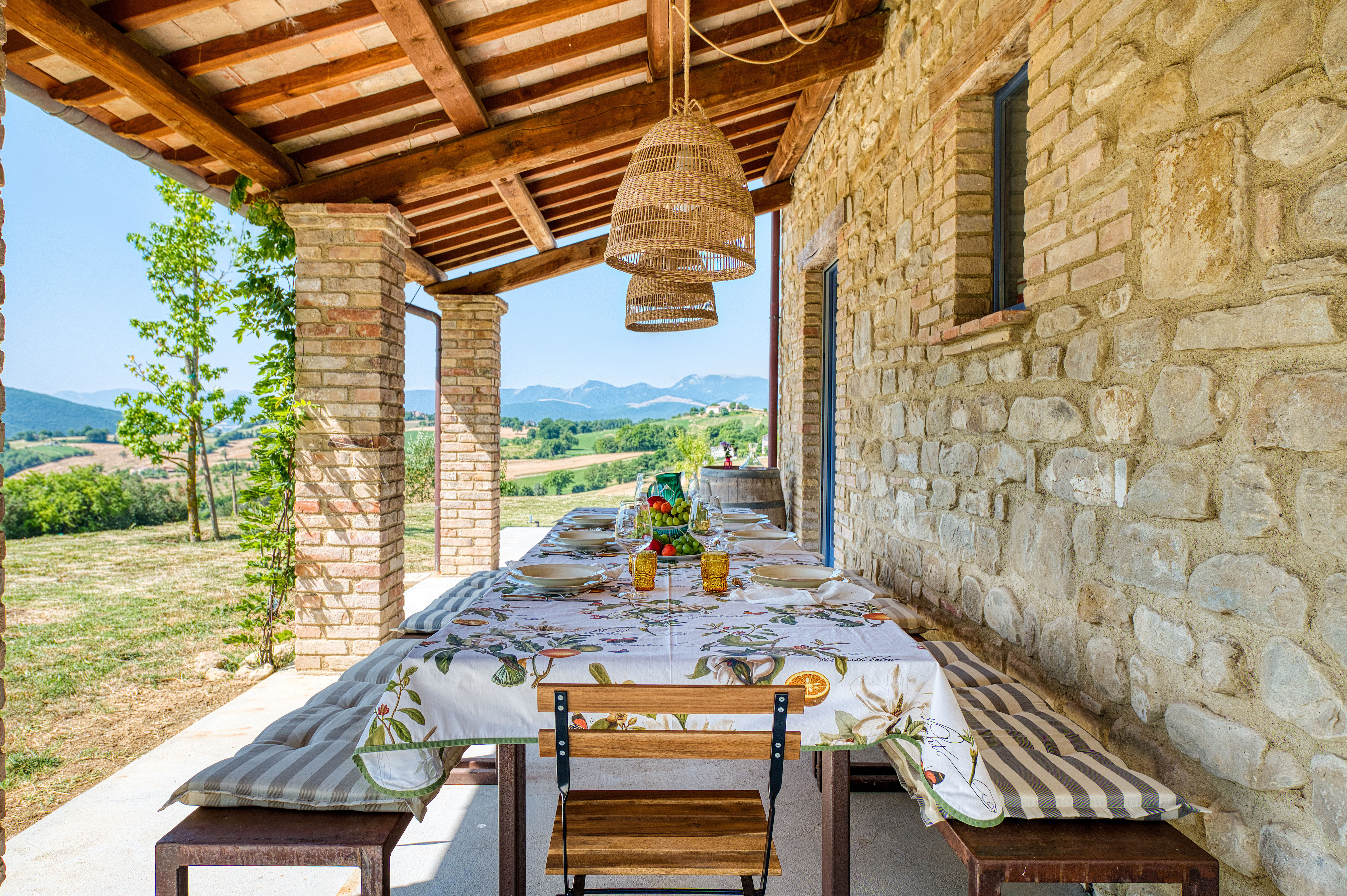A long stone farmhouse table set with striped linens and citrus sits beneath a wood-beamed stone portico overlooking a countryside view.