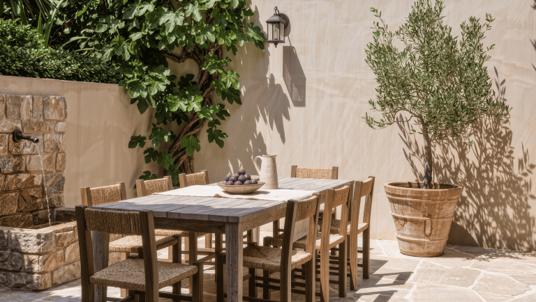A restrained European courtyard with limestone pavers, a weathered teak table, an olive tree in terracotta, and a stone wall fountain at midday.