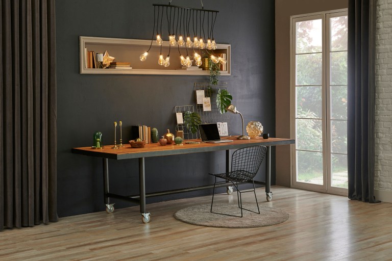 A wood desk with a laptop and brass accents set against a charcoal wall, framed by floor-length curtains and a tall window in a styled home-office nook.