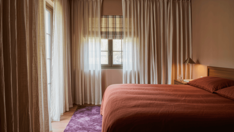 A bedroom with floor-to-ceiling oatmeal linen curtains layered over a striped Roman shade, a walnut bed with rust linen, and diffuse daylight.
