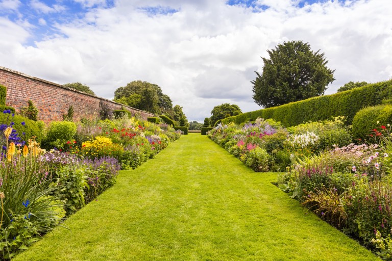A long curved herbaceous border of layered summer perennials runs beside a mown lawn inside a walled English garden.