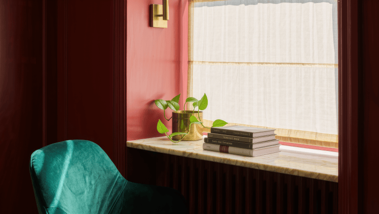A deep marble window recess with a sheer curtain diffuses bright daylight across a small brass planter and stacked books.