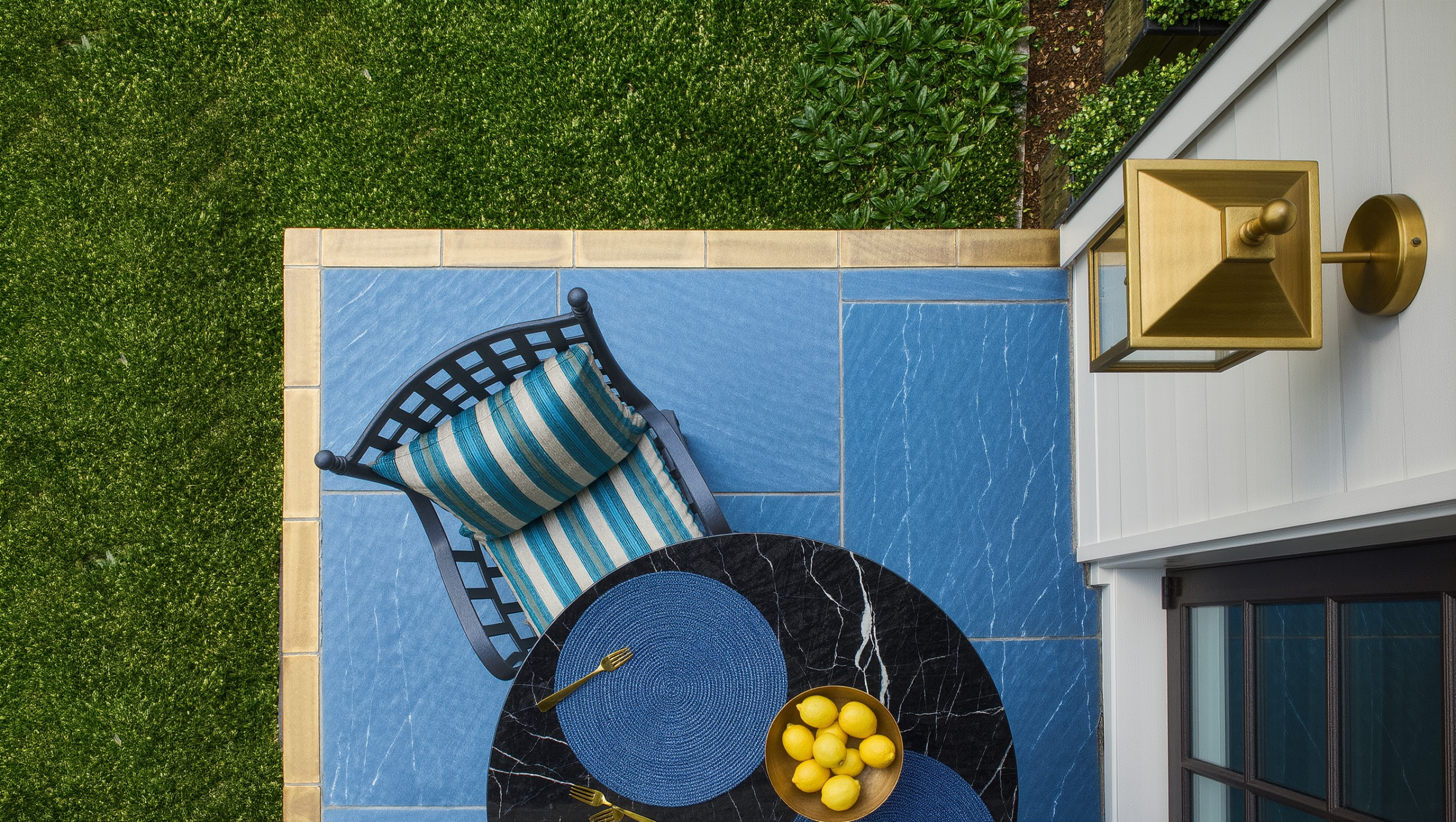 A black-marble bistro table on a bluestone dining terrace at the edge where it meets a trimmed lawn, marked by a low brass border strip.