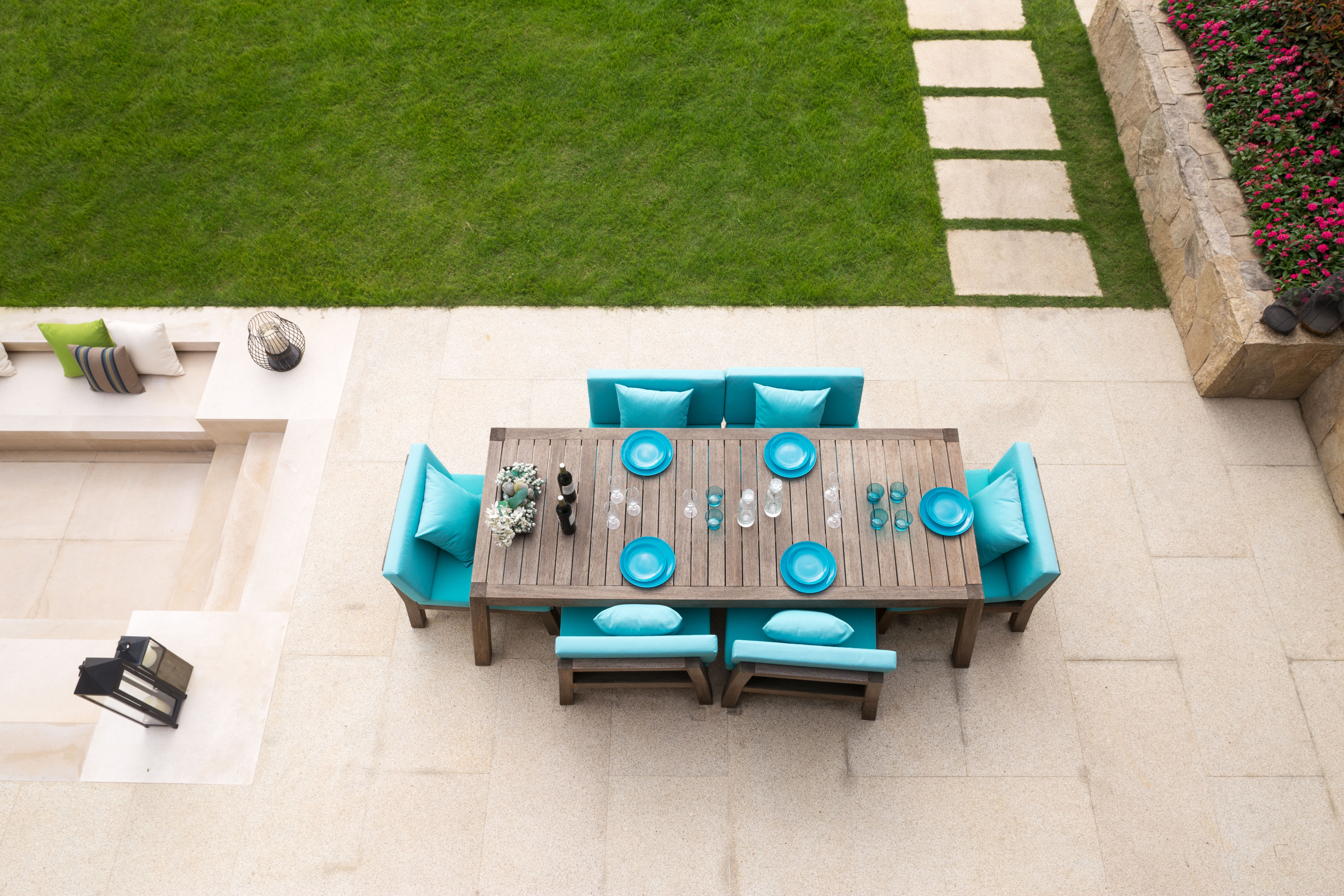 An aerial view of a stone dining terrace set with a teak table and turquoise cushions, opening directly onto a manicured lawn edged with stepping stones.