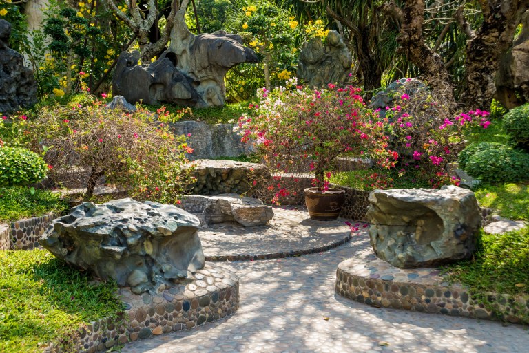 A richly planted stone garden courtyard with curved boulder seating, a pebble path, and flowering shrubs in bloom, its irrigation kept entirely out of sight.