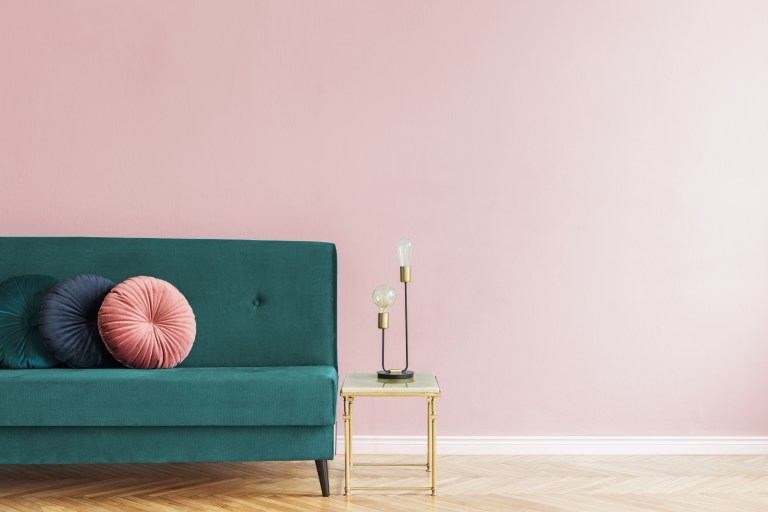 A tufted emerald velvet sofa with a round blush pillow sits against a soft pink wall beside a slim brass-legged side table.