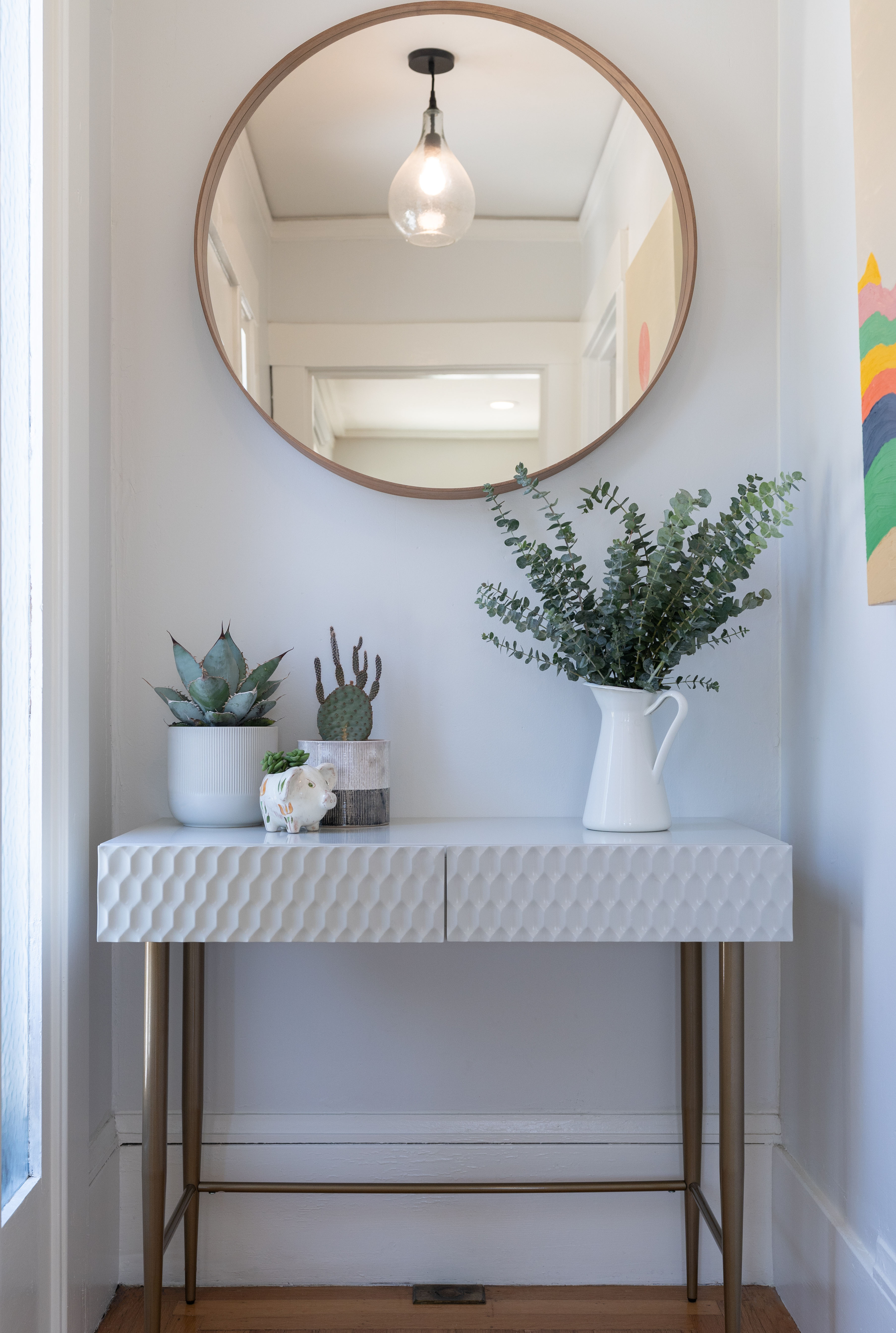 A slim marble-topped console table on brass legs stands beneath a round brass-framed mirror, styled with potted greenery and small ceramics in a bright entry hall.
