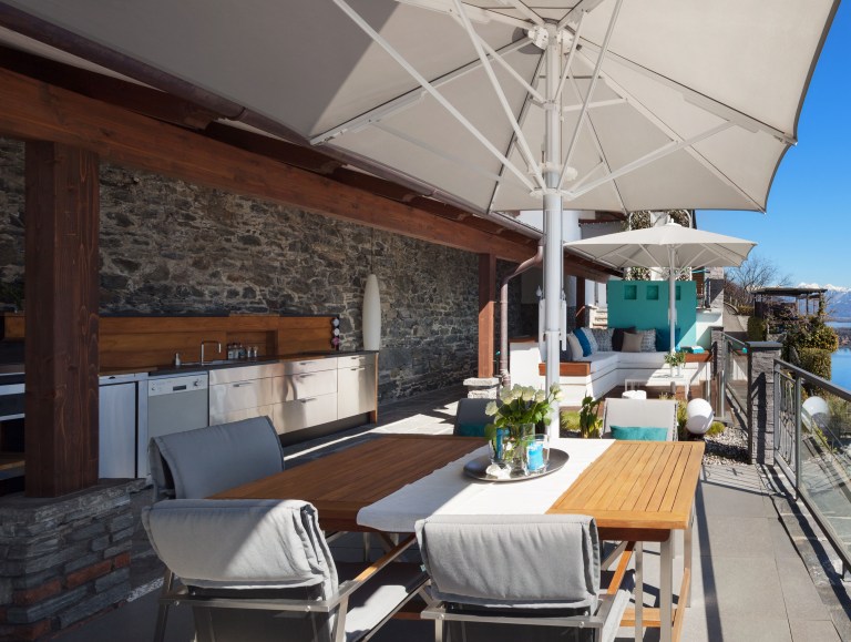 A sunlit rooftop terrace with a wood dining table set under a large umbrella, a built-in outdoor kitchen along a stone wall, and a distant sea view.