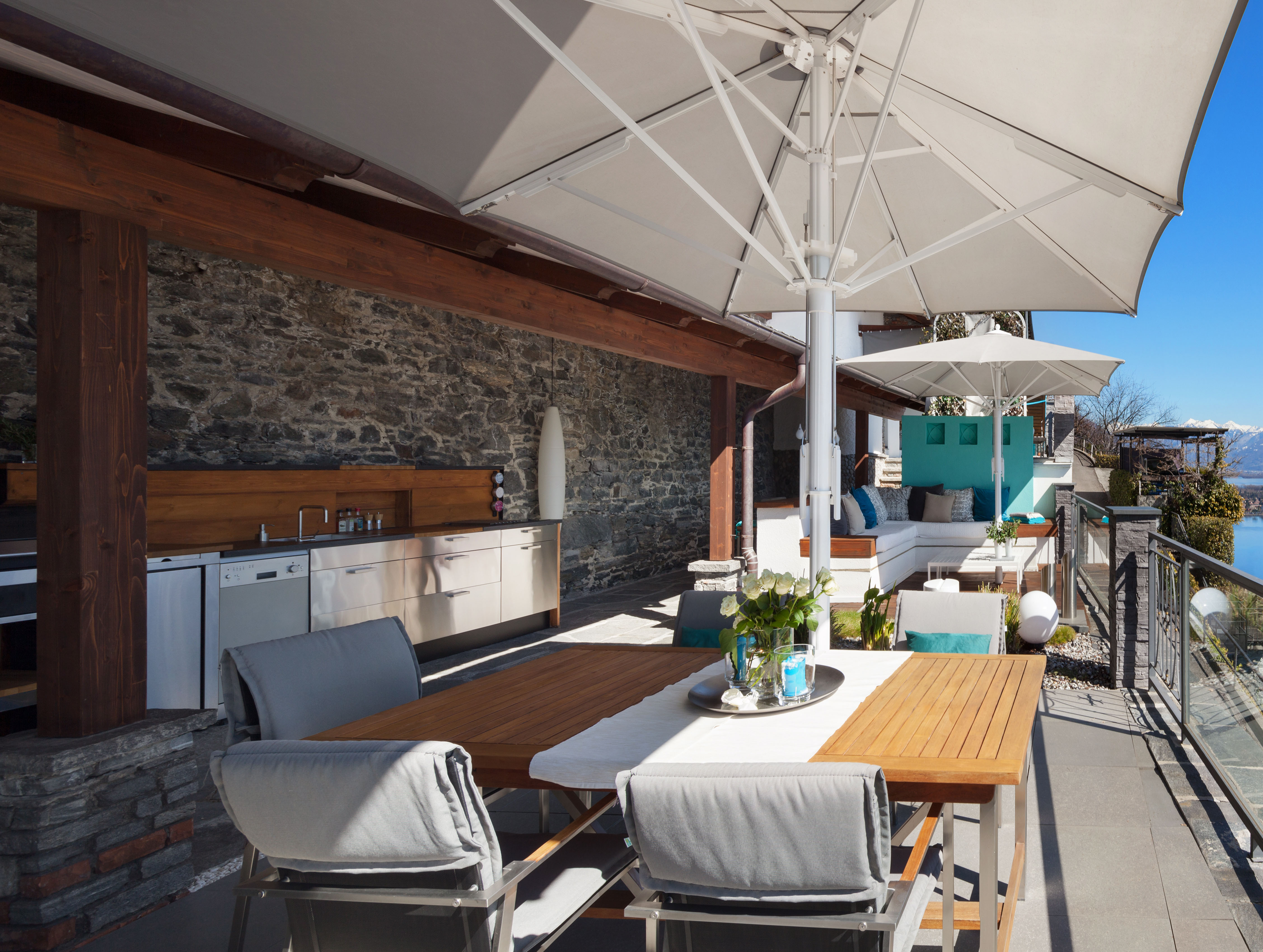 A sunlit rooftop terrace with a wood dining table set under a large umbrella, a built-in outdoor kitchen along a stone wall, and a distant sea view.