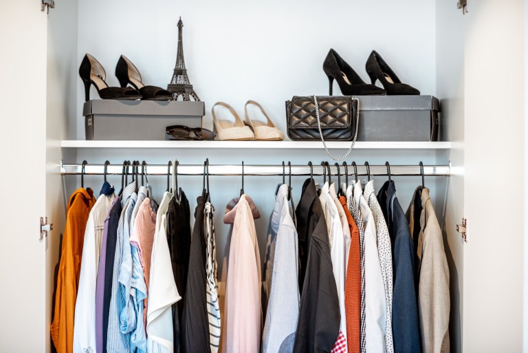 A closet rod holds a full row of hanging clothes in mixed colors, with handbags and heels lined up on the shelf above.