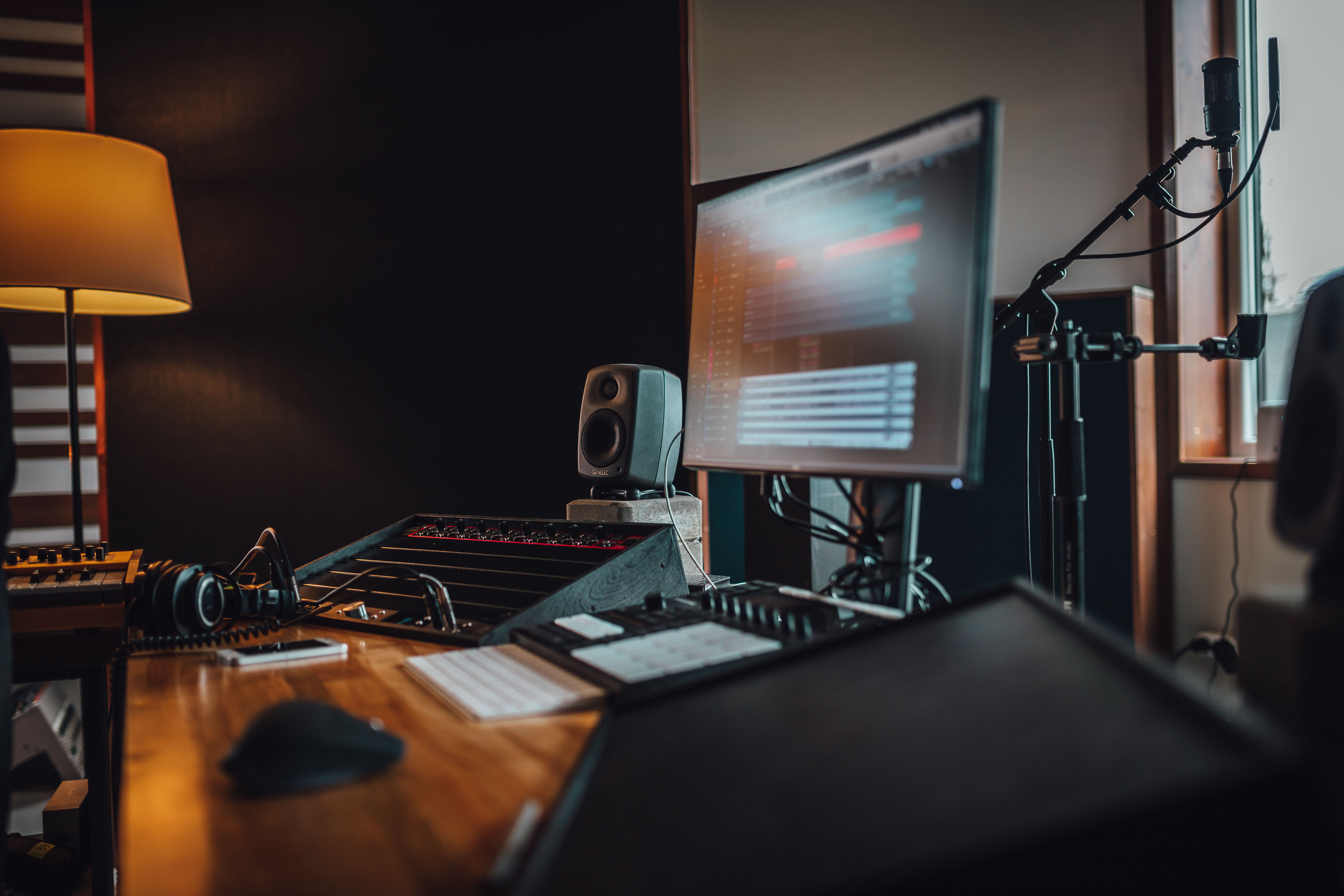 A warmly lit home recording corner with dual monitors, a synth keyboard, and a studio microphone against a dark accent wall.
