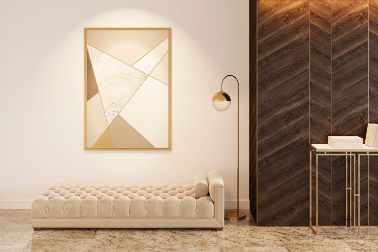 A large gold-framed abstract artwork anchors the wall above a tufted cream bench, beside dark wood paneling and a marble-topped brass console table.