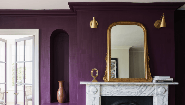 A gilt-framed mirror centered above a marble mantel on a deep plum chimney breast, with a brass sculptural object and a shadowed side alcove.