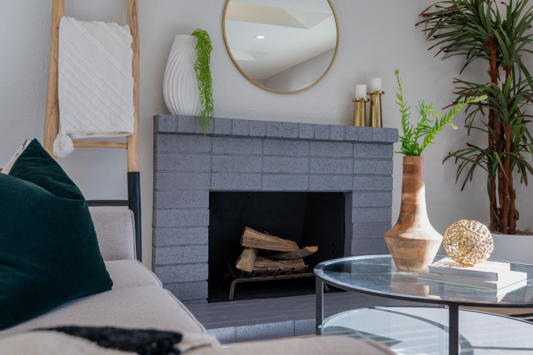 A painted brick fireplace with a white mantel, round mirror, and brass-accented styling in a light, polished living room.