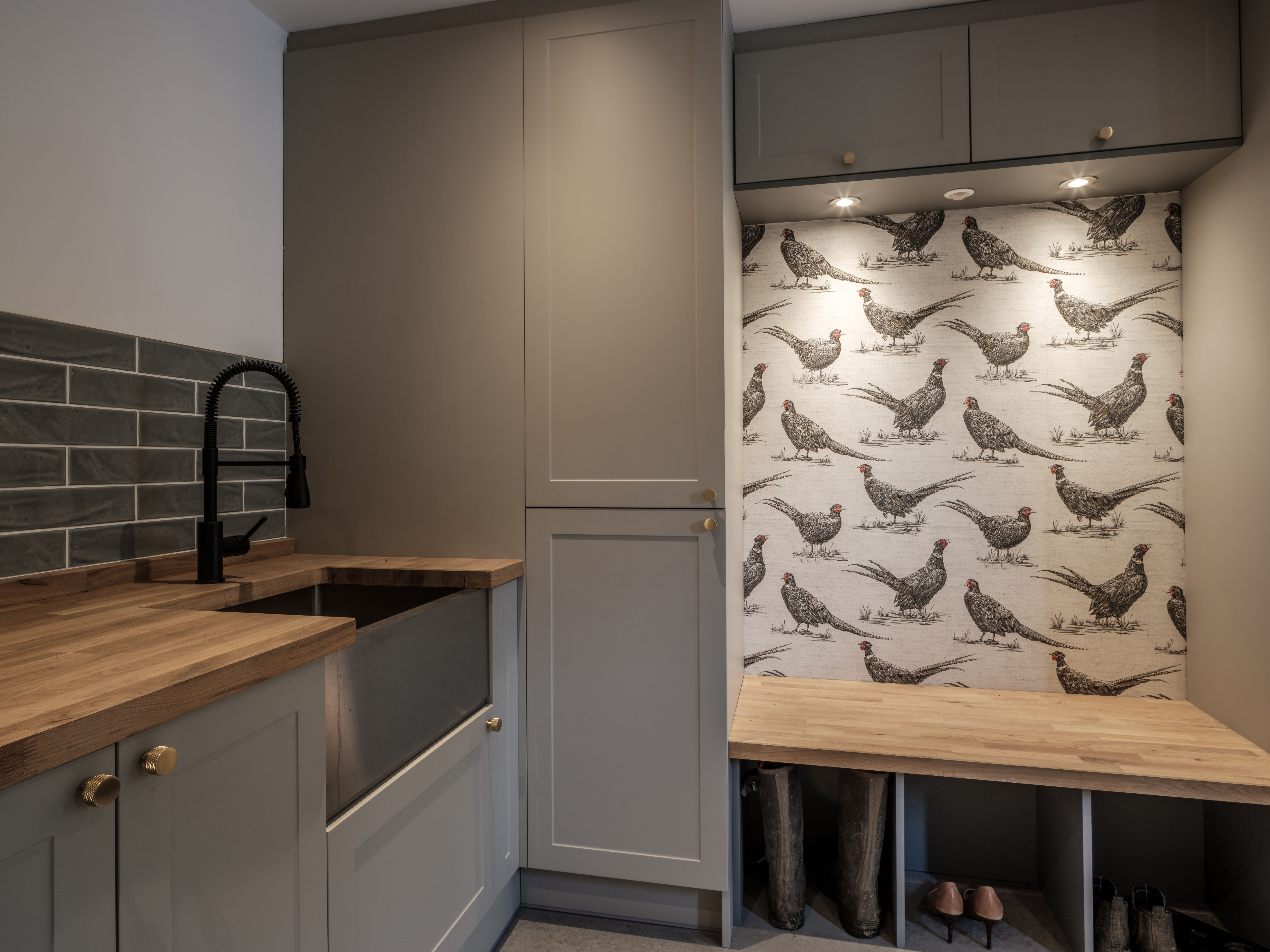 Sage-gray laundry cabinetry and a butcher-block counter beside a mudroom bench with pheasant-print wallpaper and brass hooks.